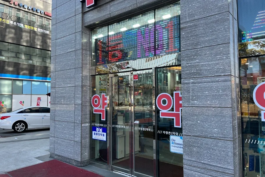 약국 출입문 측면에서 본 통유리 시공 부위, 상단 전광판과 필름이 시공된 유리문