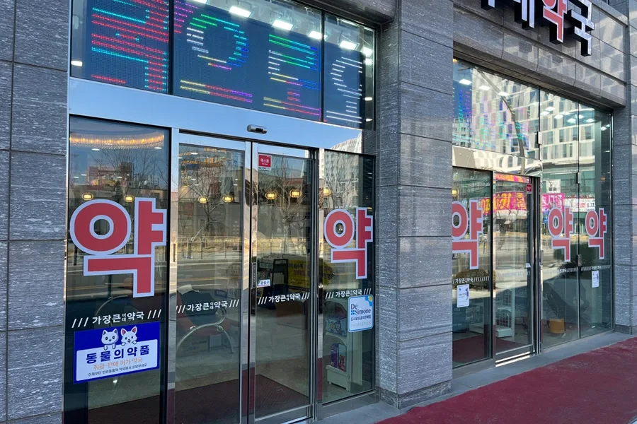 약국 정면 출입문 유리에 필름을 시공한 모습, 약 로고 스티커와 전광판이 함께 보이는 매장 입구