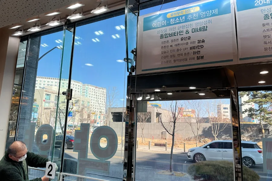 약국 내부 직원이 통유리 안쪽에서 응대하는 모습, 시공된 유리문 너머로 도로가 보이는 매장 내부