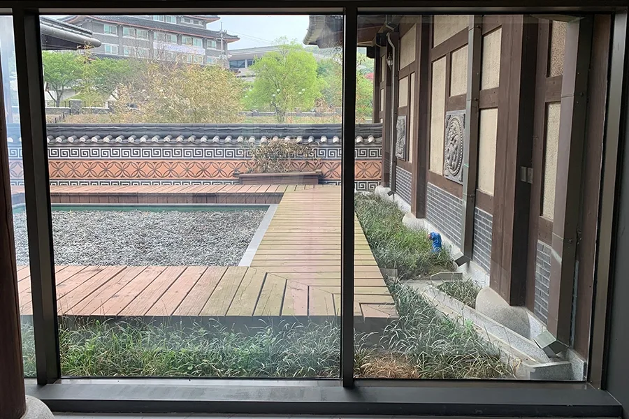 통유리 너머로 보이는 마당, 자갈과 나무 데크, 전통 담장이 어우러진 풍경