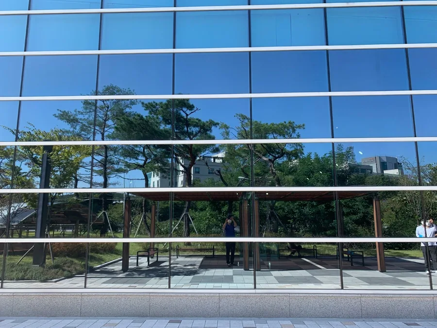 가로 프레임이 촘촘한 체육센터 로비 통유리 외벽, 야외 정자와 벤치가 유리에 반사된 모습