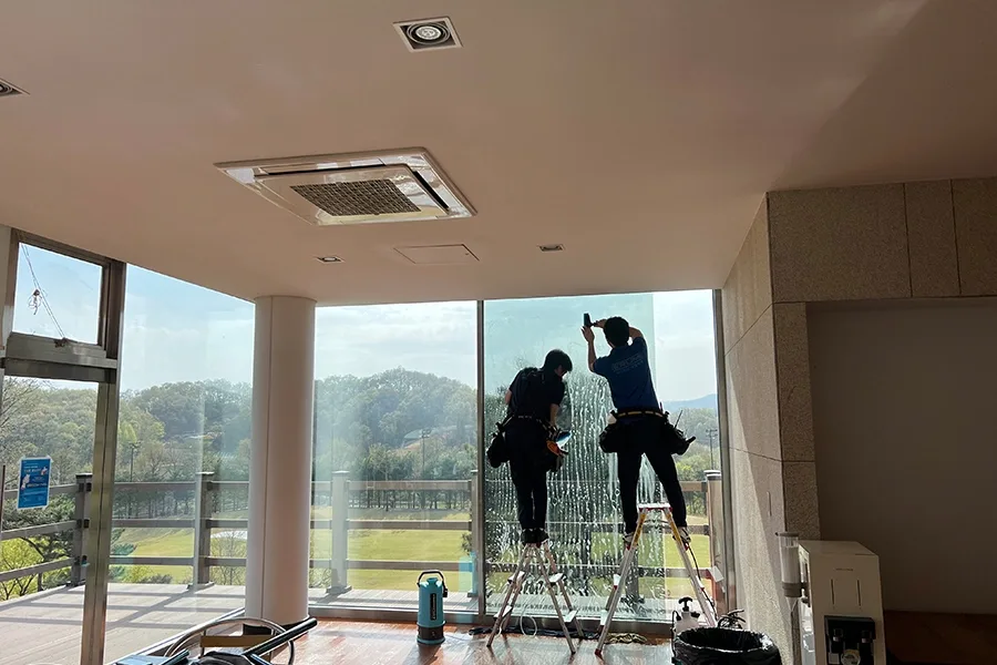 사다리 위에서 두 작업자가 통유리 상단부에 필름을 부착하는 모습, 창밖으로 잔디밭이 보임