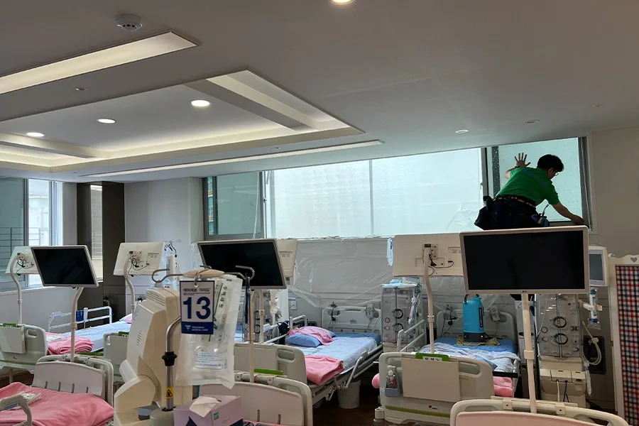 시공기사가 병상 위에서 창문 상단까지 손을 뻗어 필름을 부착하는 모습, 주변 장비는 비닐로 보호되어 있음